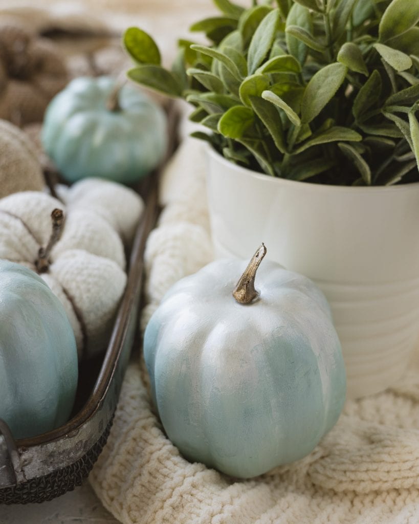A light blue faux pumpkin with a faux plant and a basketful of more pumpkins in the background.