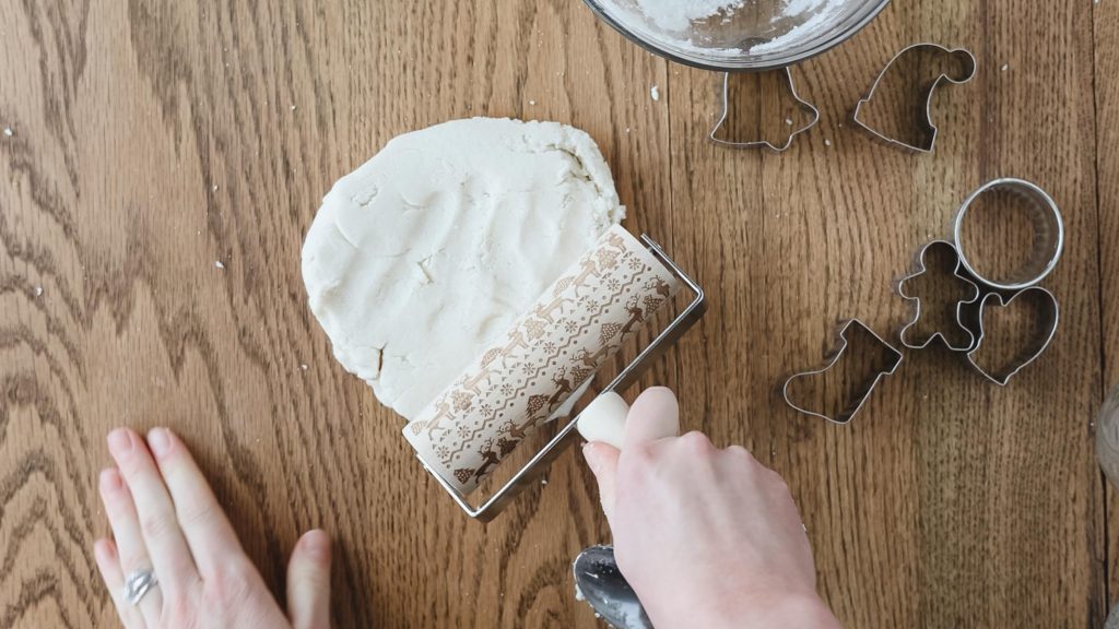 Rolling out salt dough on a wooden table with a winter-themed rolling tool.