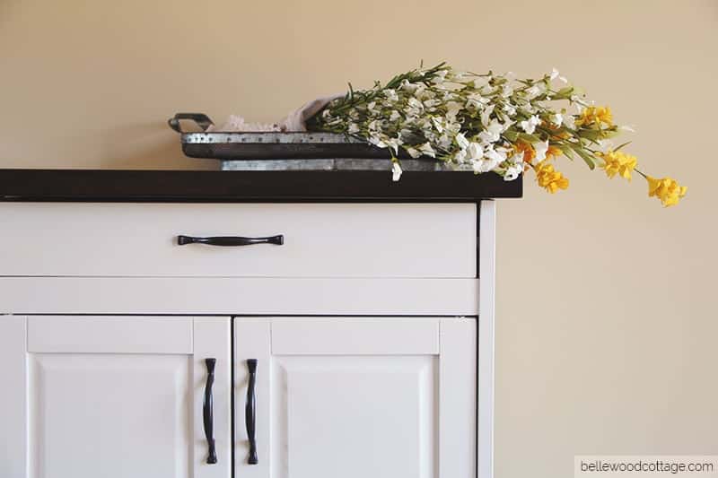 A white kitchen storage cart with a dark stained top and a basket of farmhouse flowers.