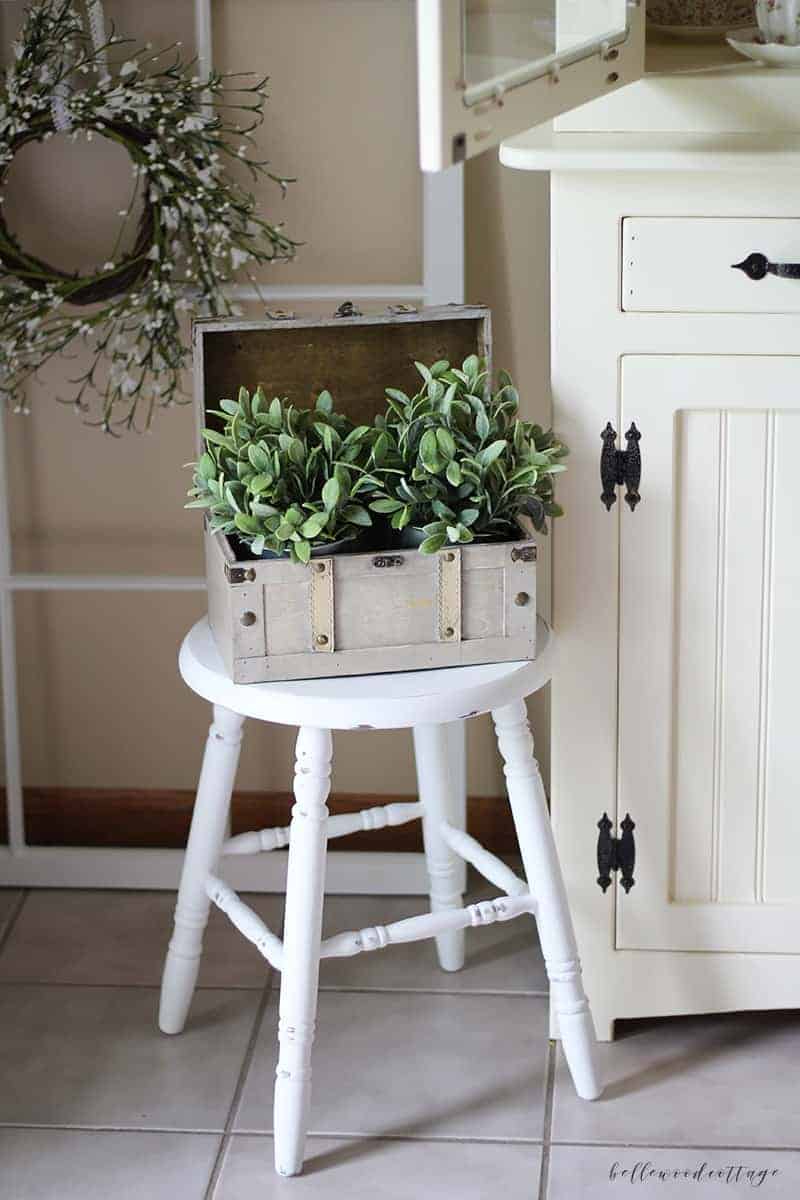 A farmhouse stool painted white and distressed with decorative greenery on top.