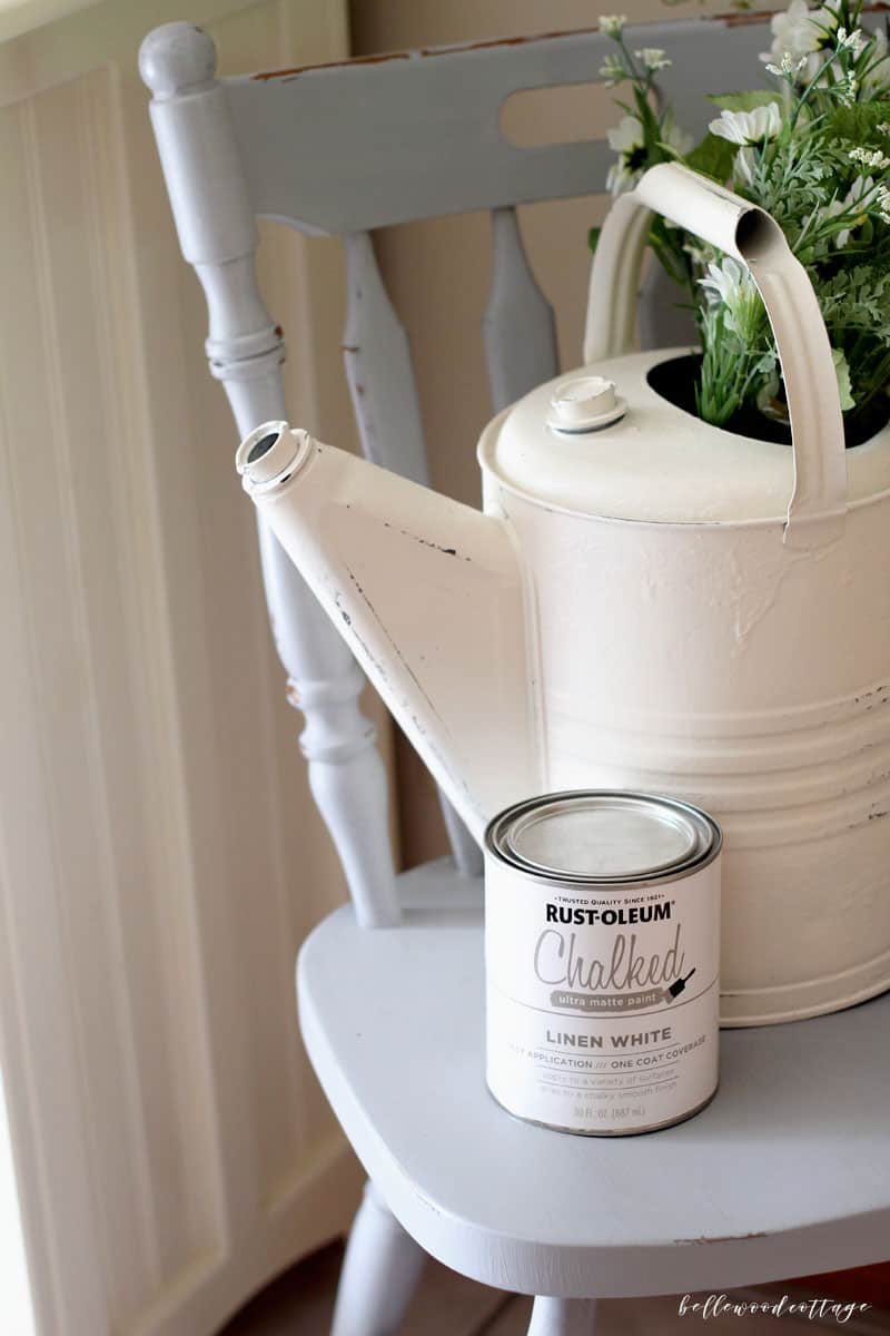 A can of chalk paint on a chair with a vintage watering can behind it.