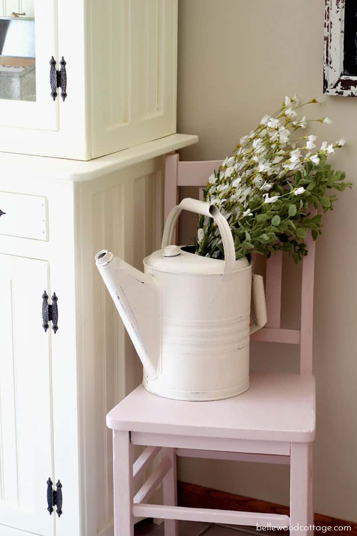 A pink farmhouse chair with a watering can full of faux flowers.