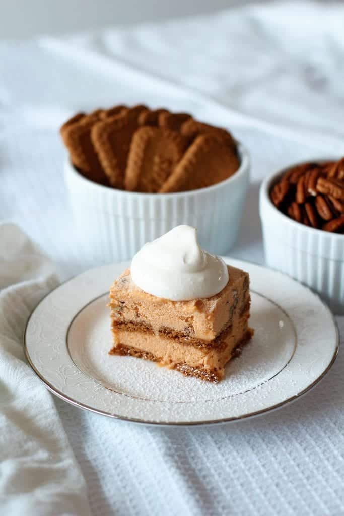 A square of Pumpkin Ice Cream Dessert on a white formal plate surrounded by ramekins of cookies and pecans.