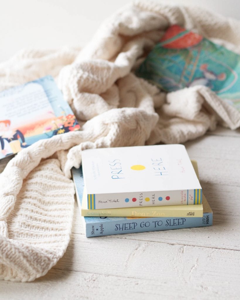 Three board books for babies on a wooden surface surrounded by a knit blanket.