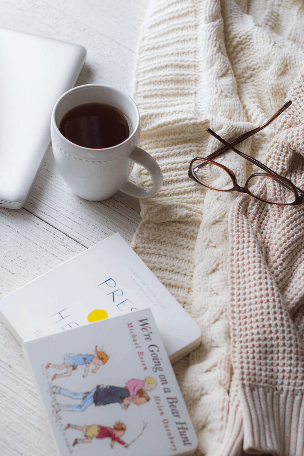 A mug of tea, board books, glasses, and a computer on a wooden surface.