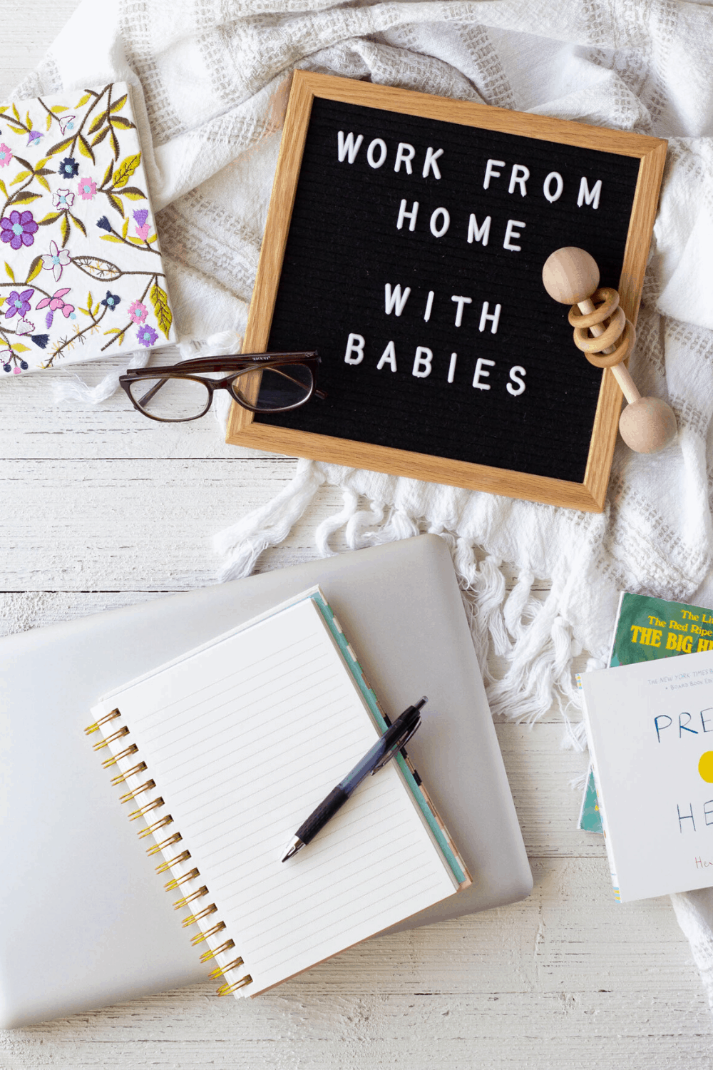 A letterboard with the words “work from home with babies”, a laptop, and notebooks on a wooden surface.