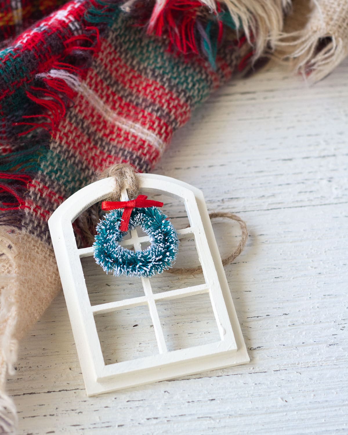 A Christmas ornament that is a window with a wreath in miniature displayed on a wooden surface.