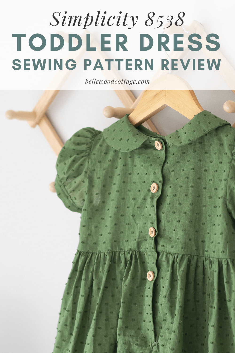 A green handmade toddler dress with wooden buttons and a Peter Pan collar hanging on a wooden hanger.