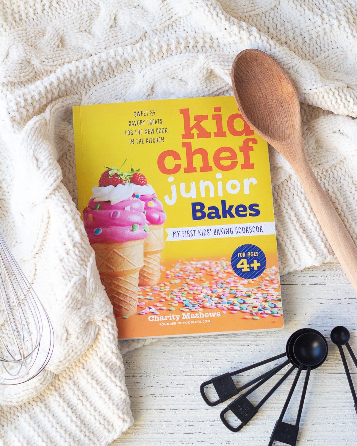 A kids baking book entitled, Kid Chef Junior Bakes, on a wooden surface surrounded by baking tools.