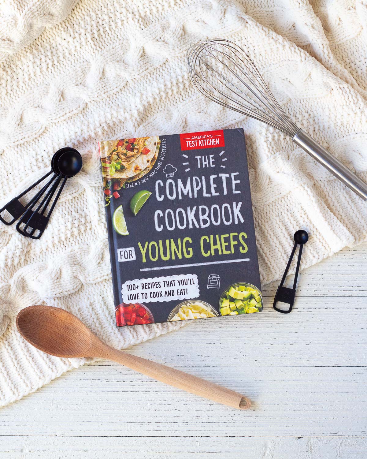 A children's cookbook on a wooden surface surrounded by cooking utensils.