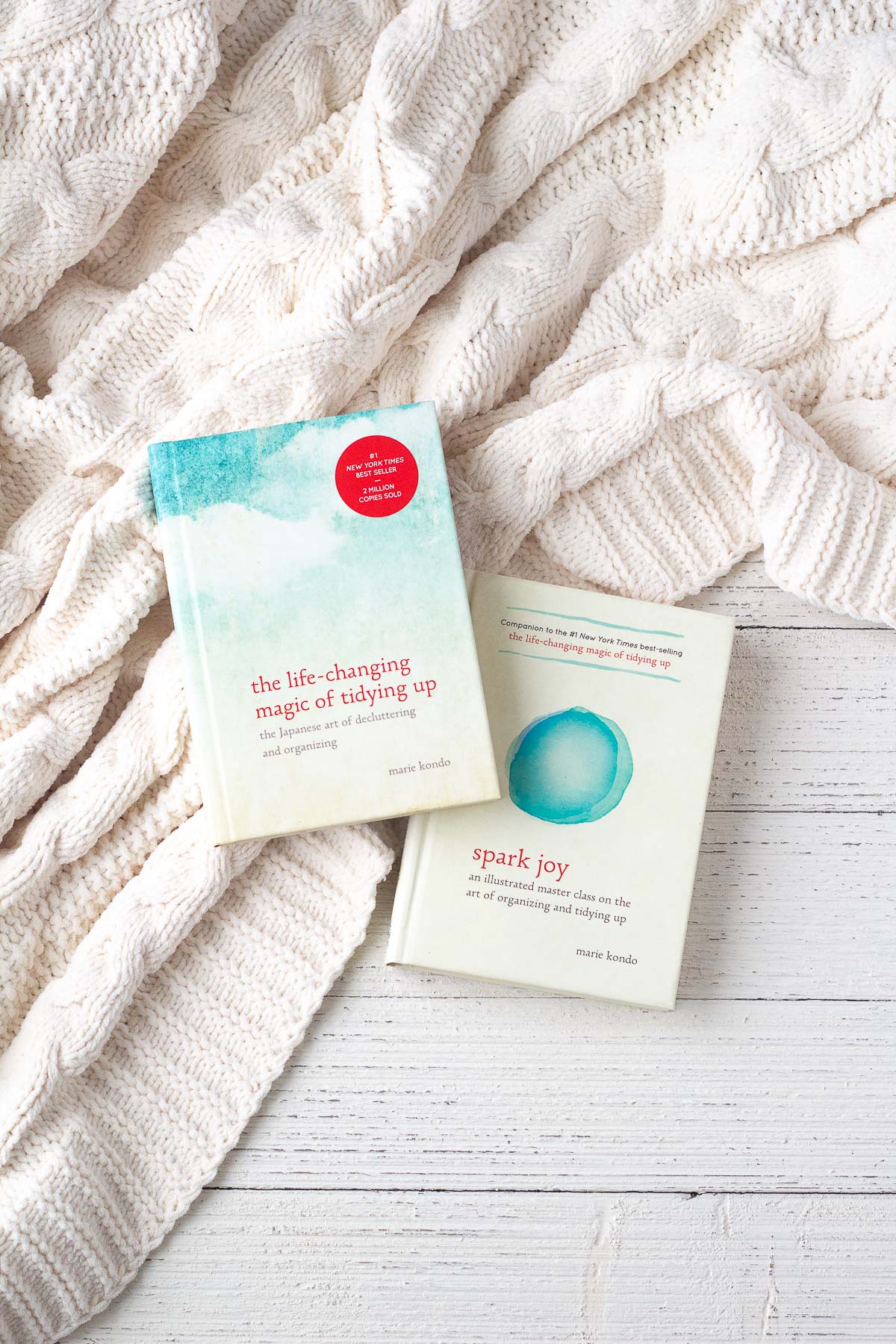 Two Marie Kondo books (which provide decluttering tips) on a knit blanket and white wooden surface.