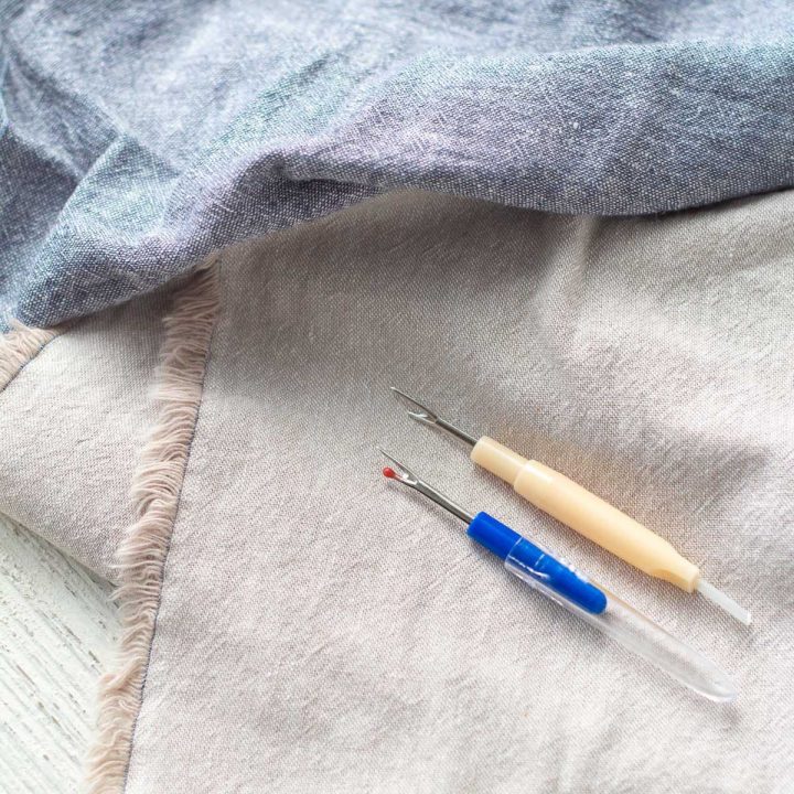 Two seam rippers on top of linen fabric.
