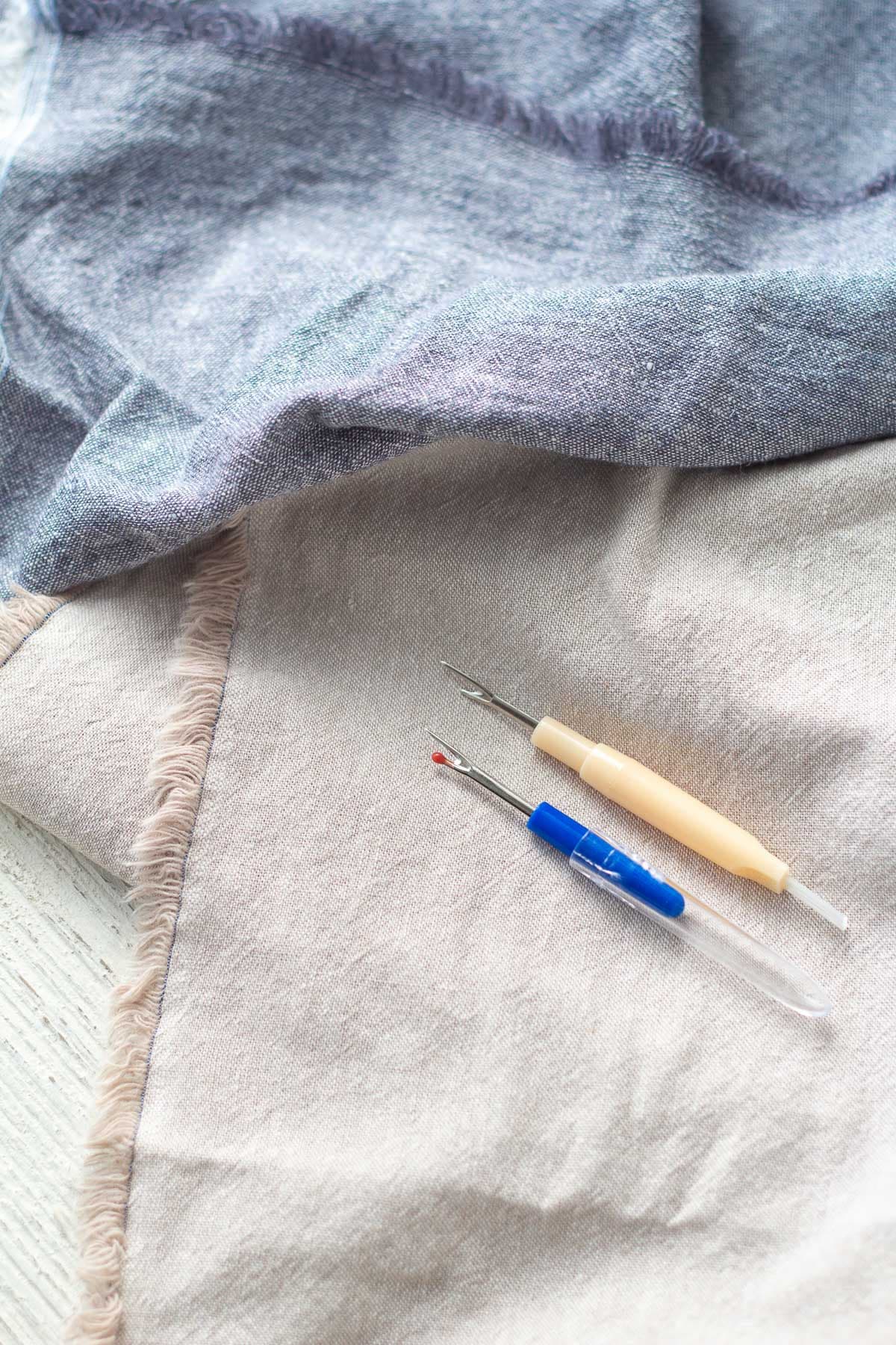 Two seam rippers on top of linen fabric.