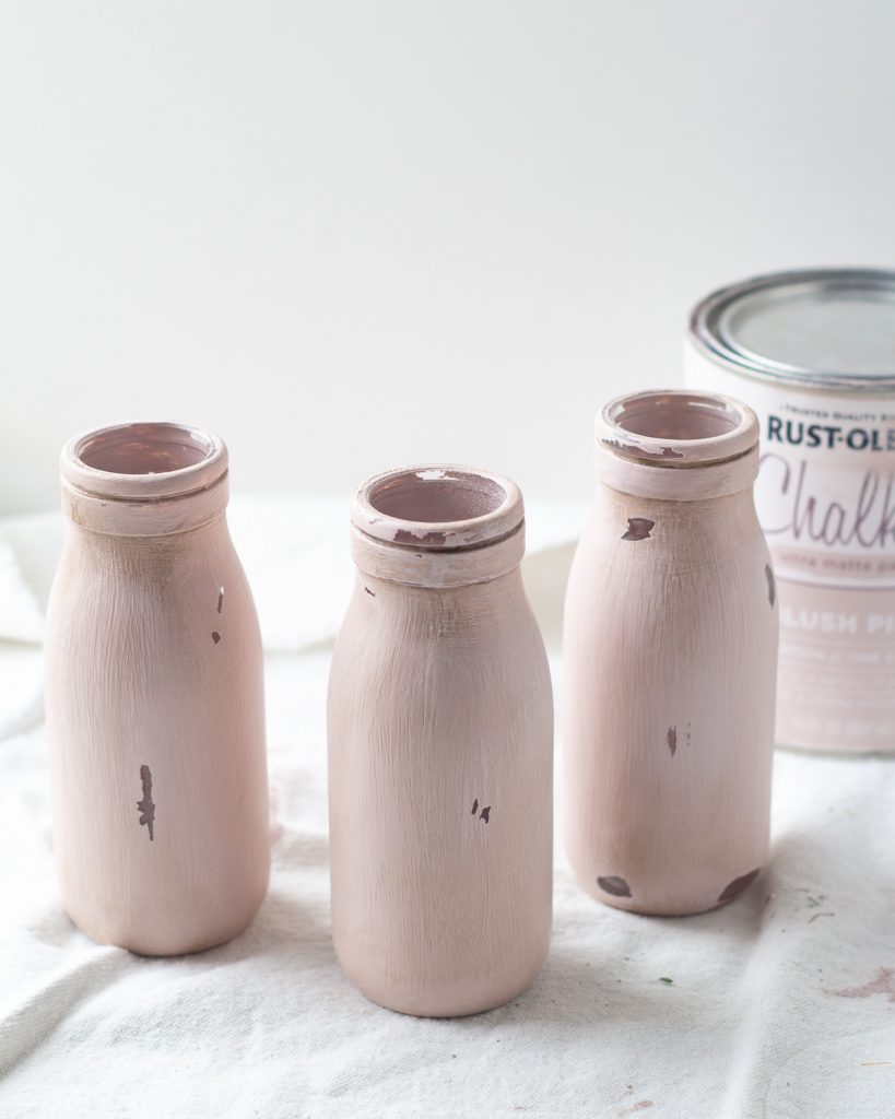 Three chalk painted and distressed milk bottles with a can of pink paint nearby.