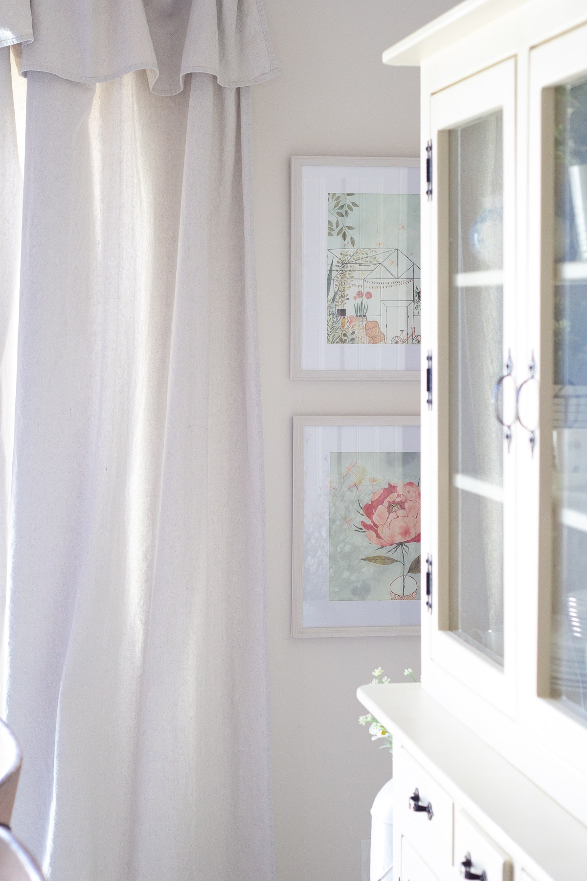 Two framed pictures on a white wall surrounded by drop cloth curtains and a white hutch.