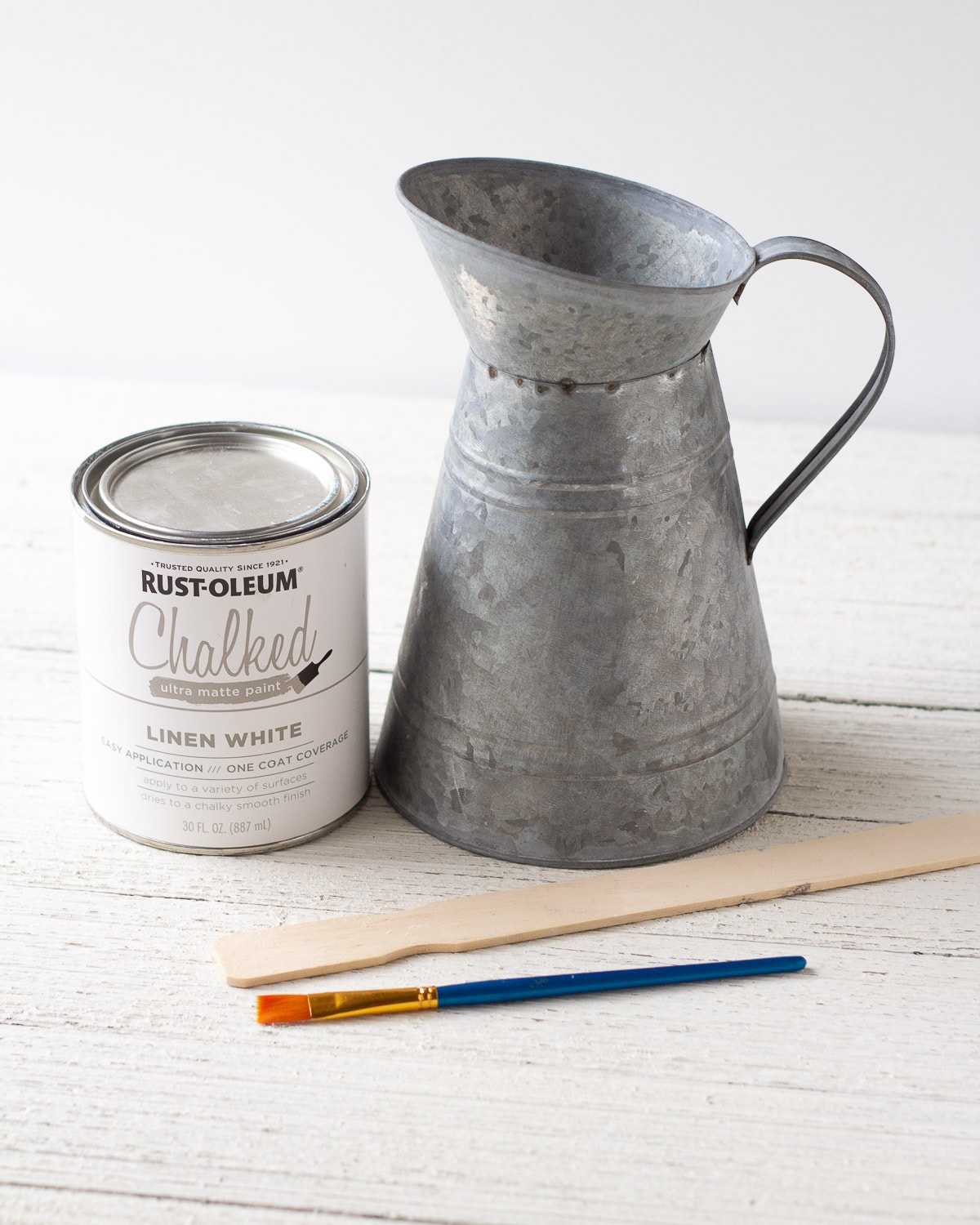 A galvanized pitcher, can of white chalk paint, paint stir stick, and a paintbrush.