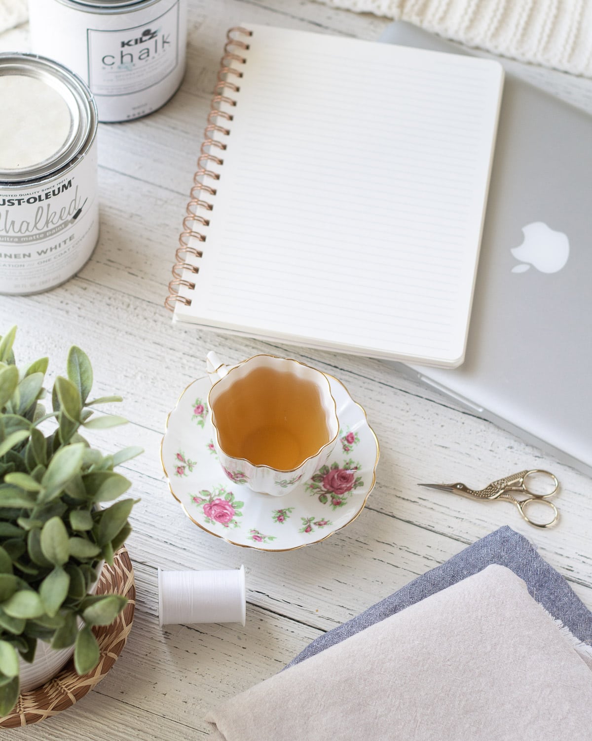 A notebook, teacup, thread, fabric, and embroidery scissors on a wooden surface.