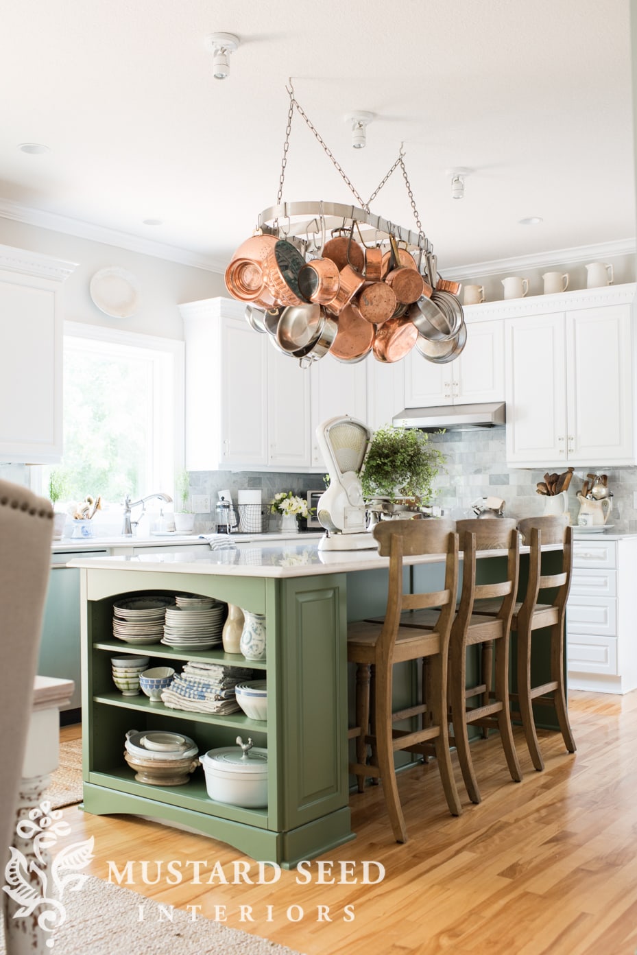 A farmhouse kitchen featuring a green island, white cabinets, and a pot rack with copper pots.