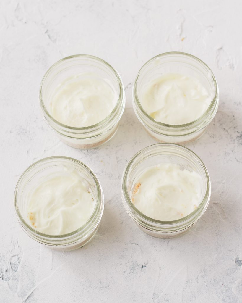 Four 4-oz mason jars with a layer of cream cheese filling spread over a no-bake crust.