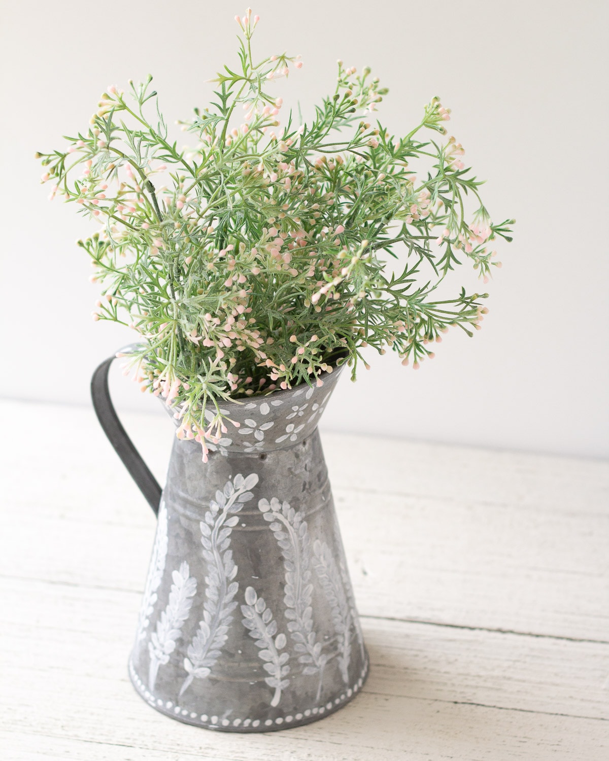 A galvanized pitcher filled with faux florals and decorated white paint floral designs.