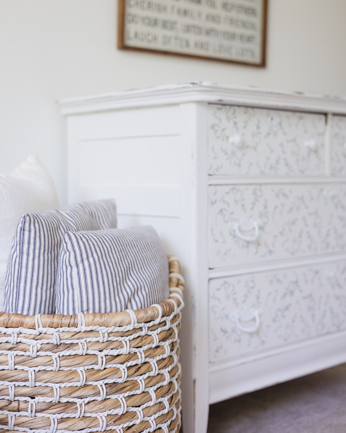 A woven basket with ticking pillows and a painted dresser in the background.