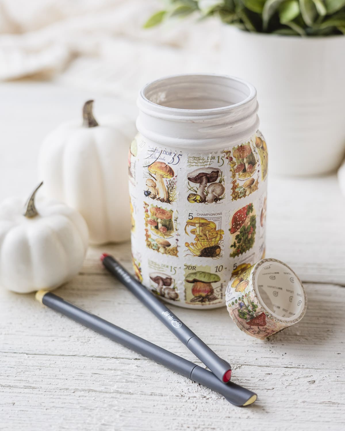 A mason jar covered with postage stamp style washi tape with faux pumpkins in the background.