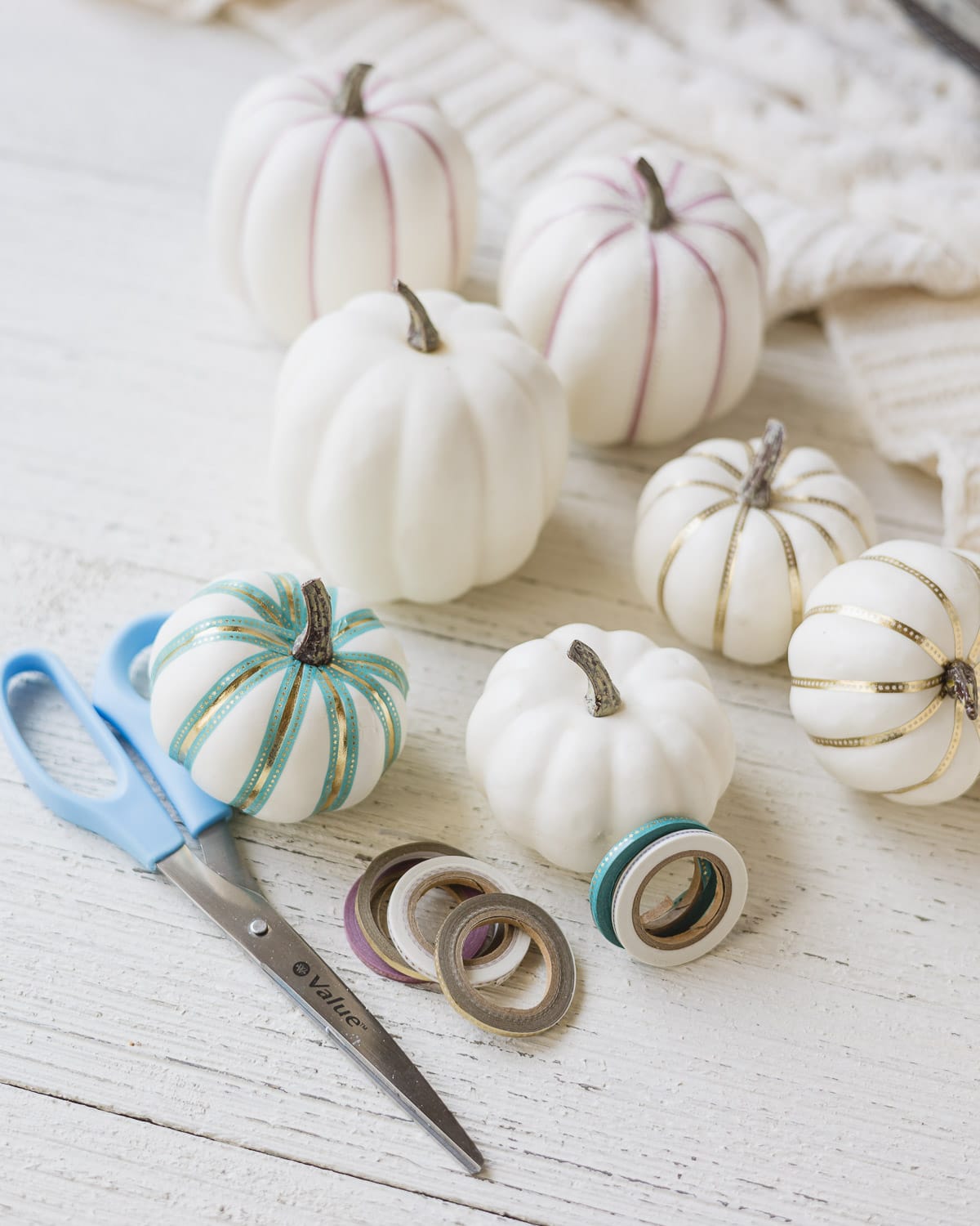 White foam pumpkins, scissors, decorative tape, and decorated pumpkins on a wooden surface.