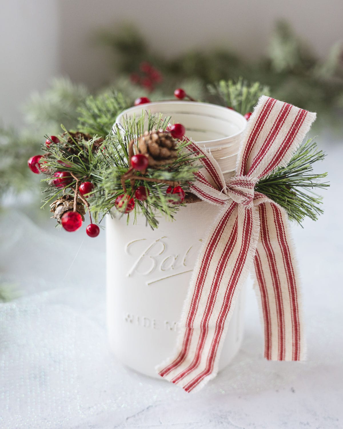 A mason jar painted white with a holiday mini wreath around the top and a red ticking stripe ribbon.