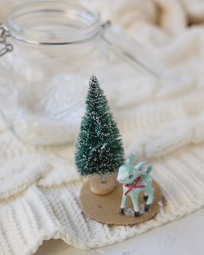 A small reindeer ornament and mini bottle brush tree on a circle of cardboard.