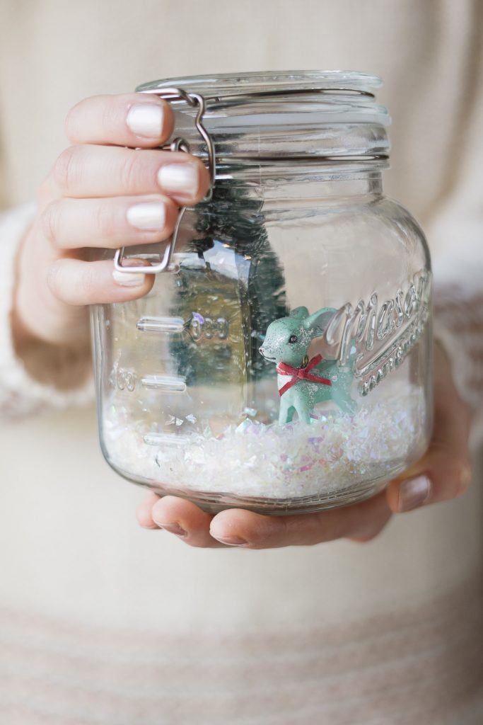 Hands holding a mason jar Christmas scene with faux snow and a mini reindeer.