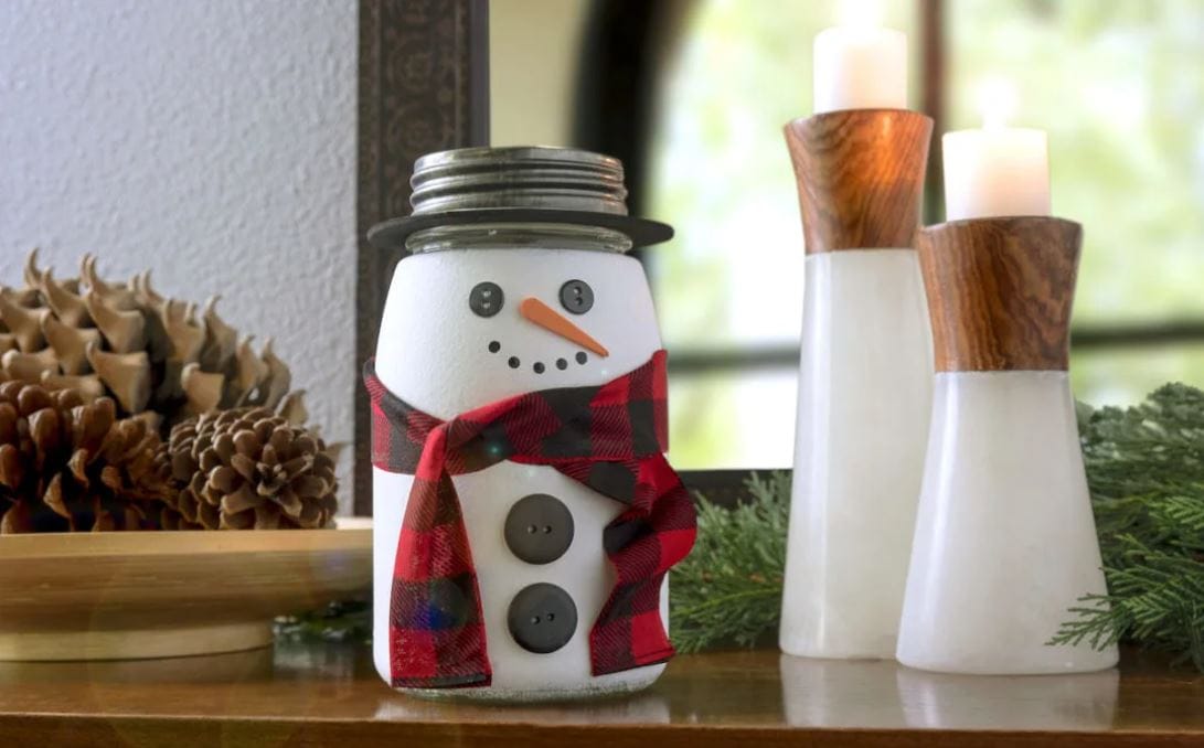 A glass jar painted as a snowman with black buttons and a plaid scarf.