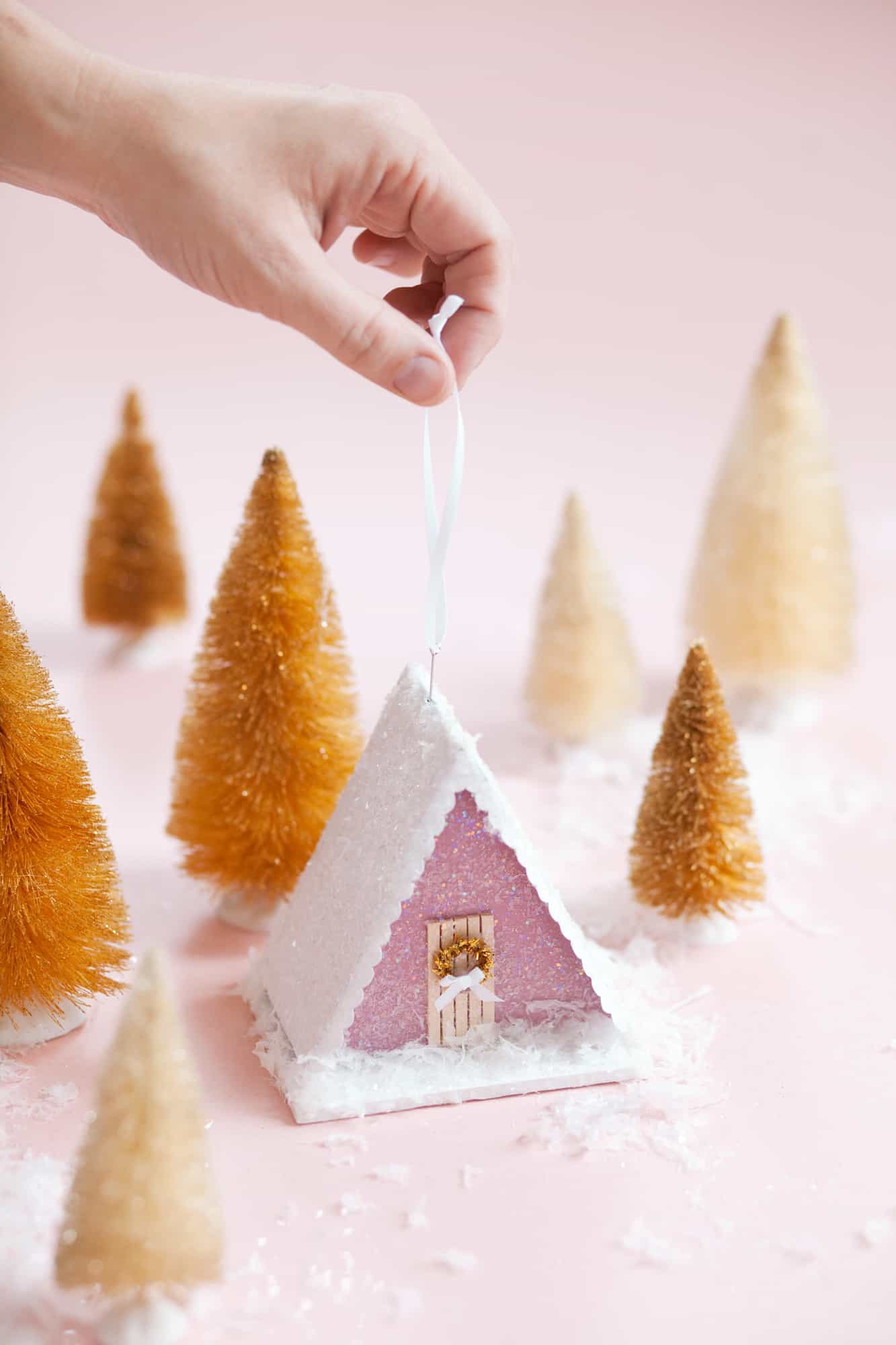 A mini pink A-frame house ornament surrounded by faux snow and bottle brush trees.