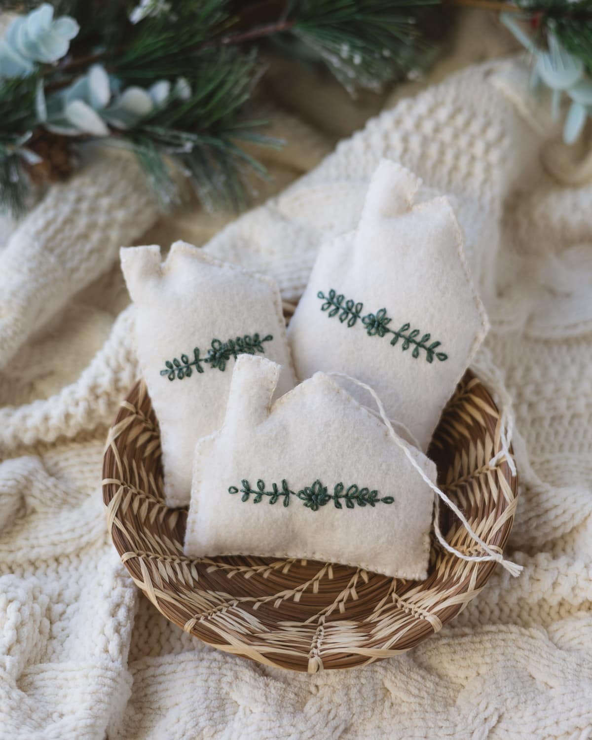 Three handmade white felt house Christmas tree ornaments in a woven basket.