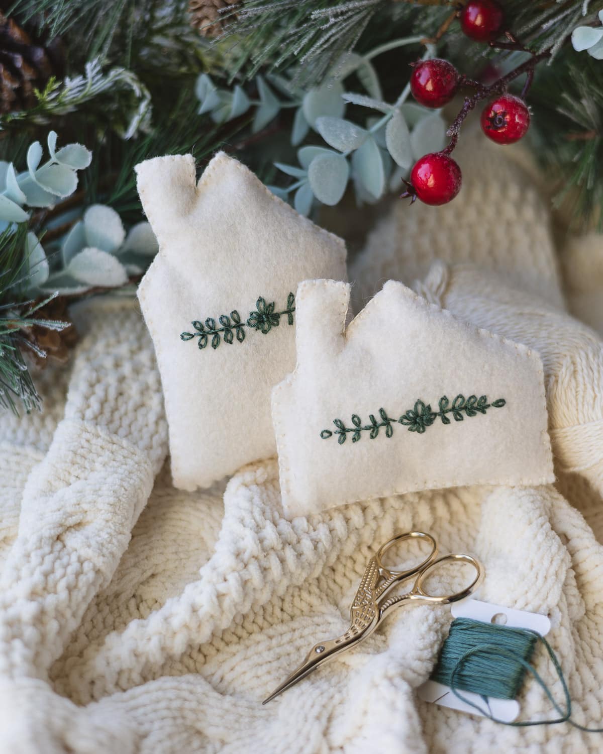 Two white felt house ornaments with a pair of gold embroidery scissors and a spool of green embroidery floss.