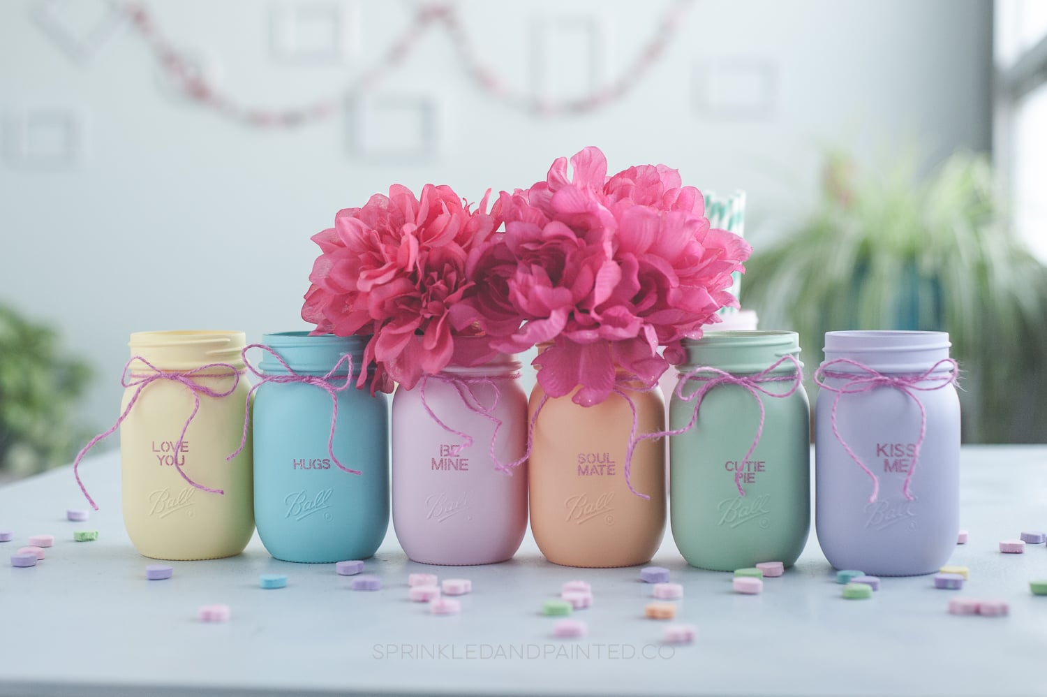 Six colorful mason jars painted in pastel candy heart colors with Valentine sayings printed on each one.