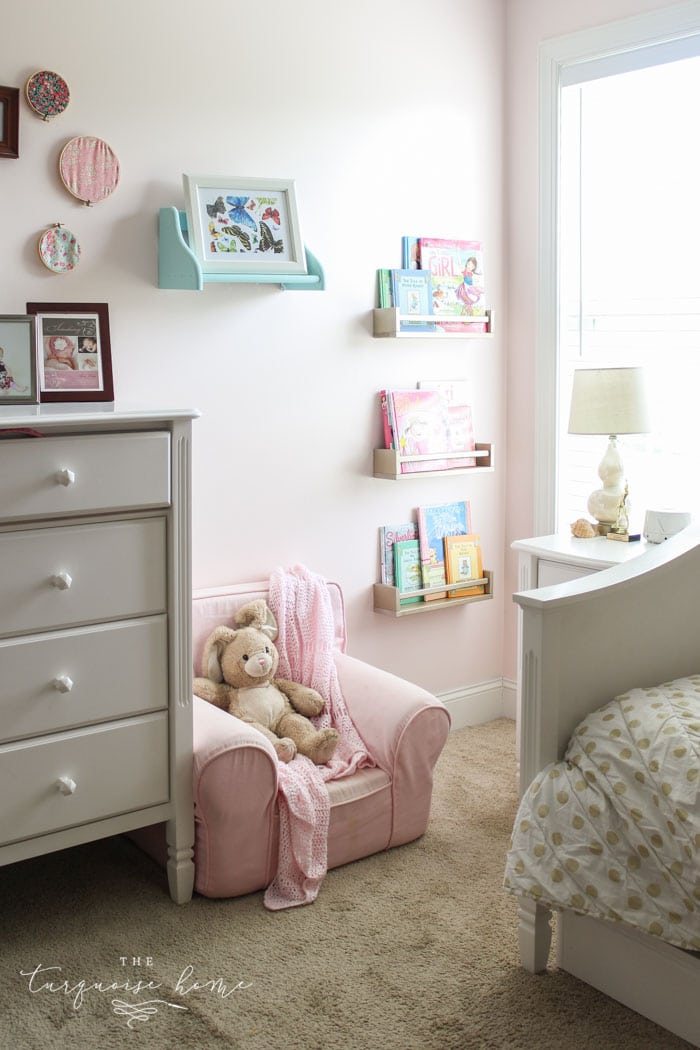 The corner of a girl's bedroom with a pink chair and bookshelves on the wall.