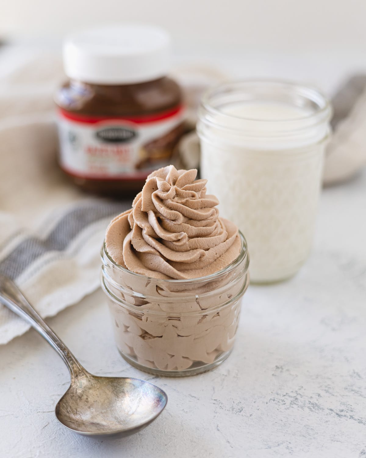 Nutella whipped cream piped into a mason with a spoon, glass of heavy cream, and a jar of chocolate-hazelnut spread in the background.