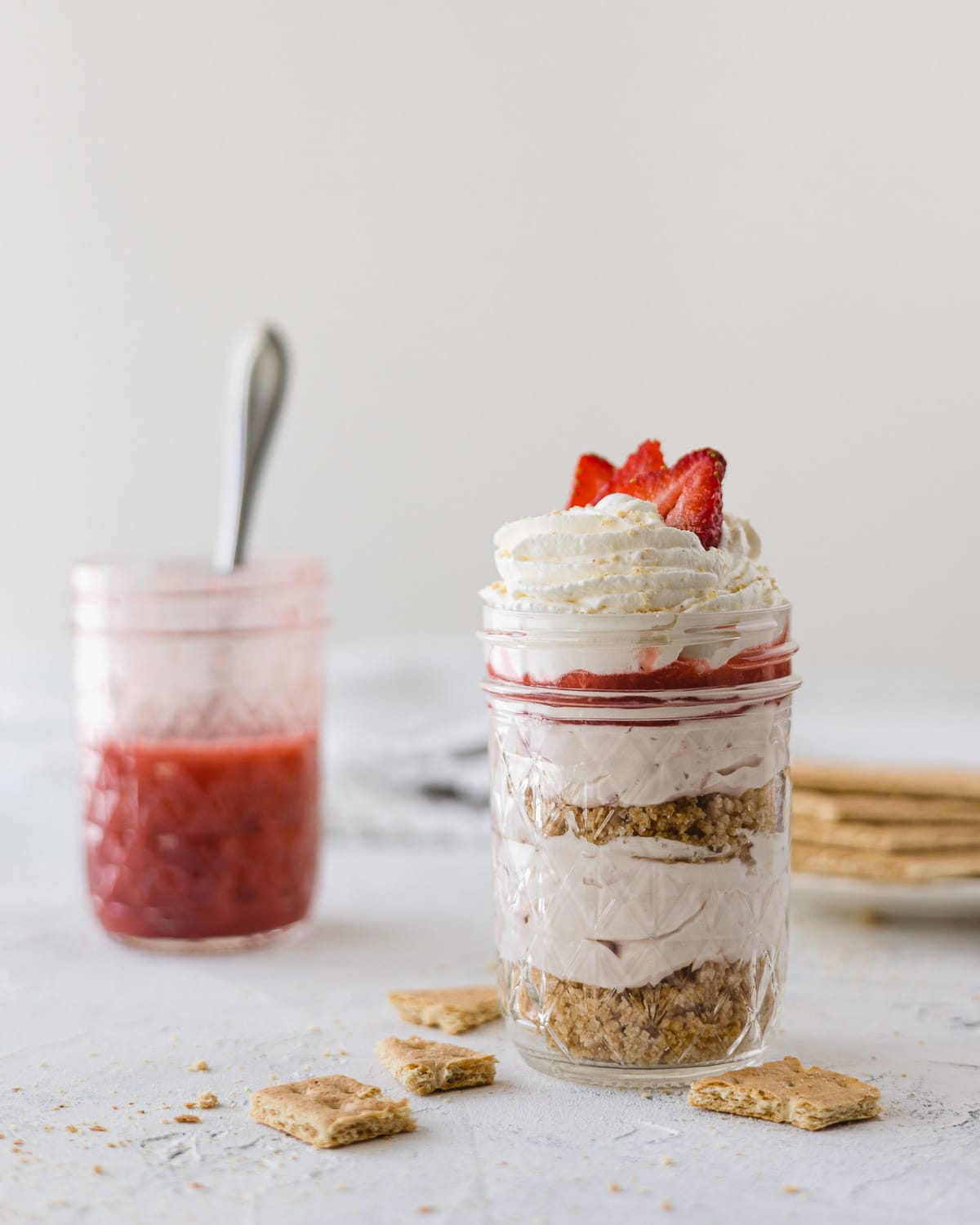 Layered no bake strawberry cheesecake in a mason jar with a jar of strawberry sauce in the background.