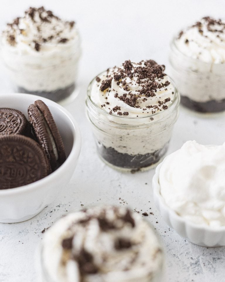Oreo mason jar desserts topped with whipped cream and cookie crumbles and a small bowl of chocolate sandwich cookies.