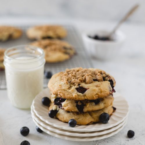 A stack of three blueberry cookies on a stack of three plates with fresh blueberries and a glass of milk.