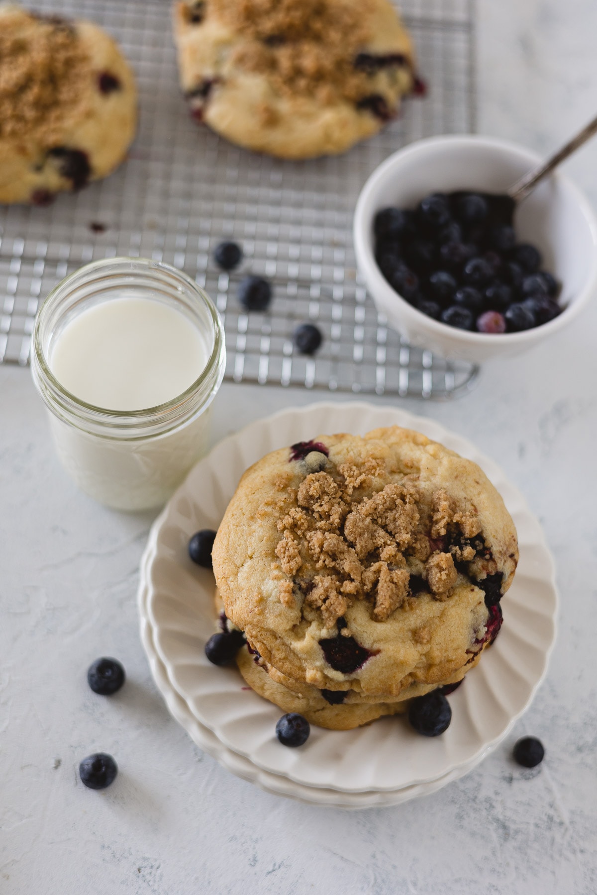 A stack of blueberry muffin cookies with streusel on white scalloped plates with fresh blueberries and a glass of milk.