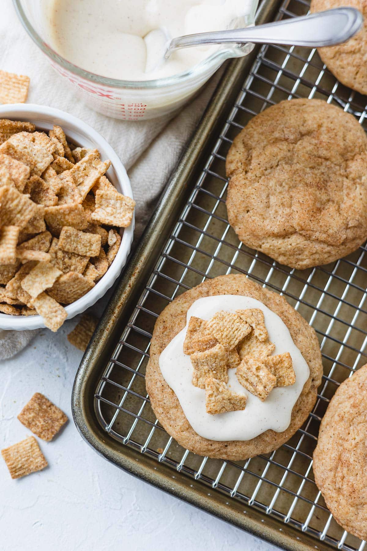 A frosted cinnamon cookie on a cookie tray and cooling rack with a sprinkle of crunchy cinnamon toast cereal on top.