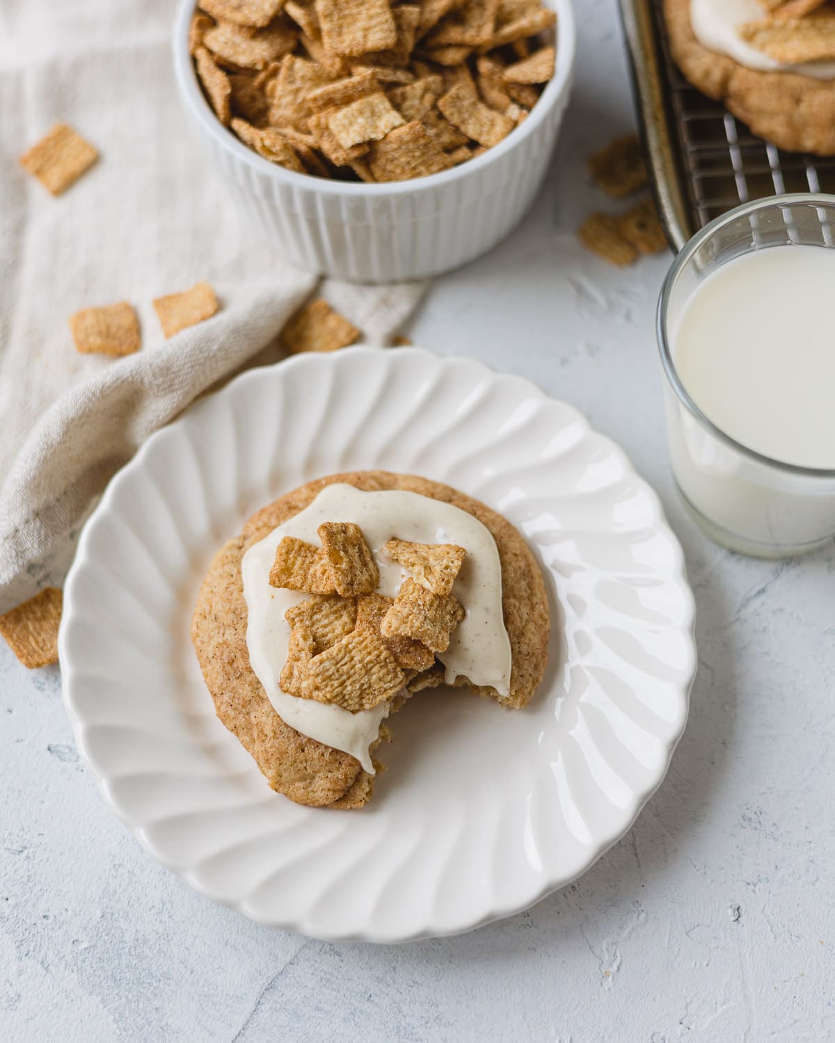 A frosted cinnamon cookie with a bite removed on a scalloped plate with a sprinkle of cinnamon cereal on top.