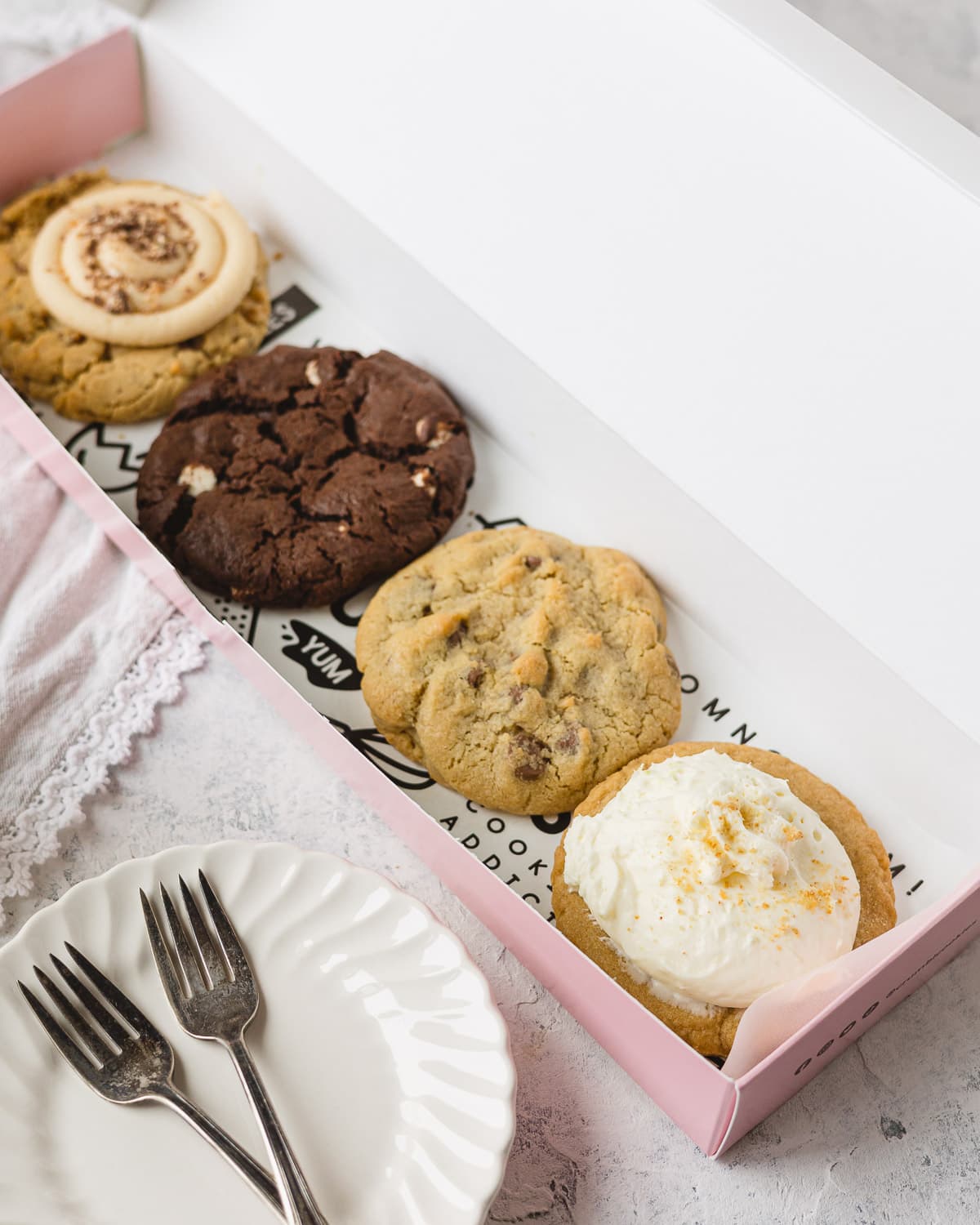 A pink box of four large bakery cookies and a stack of plates with two small forks.
