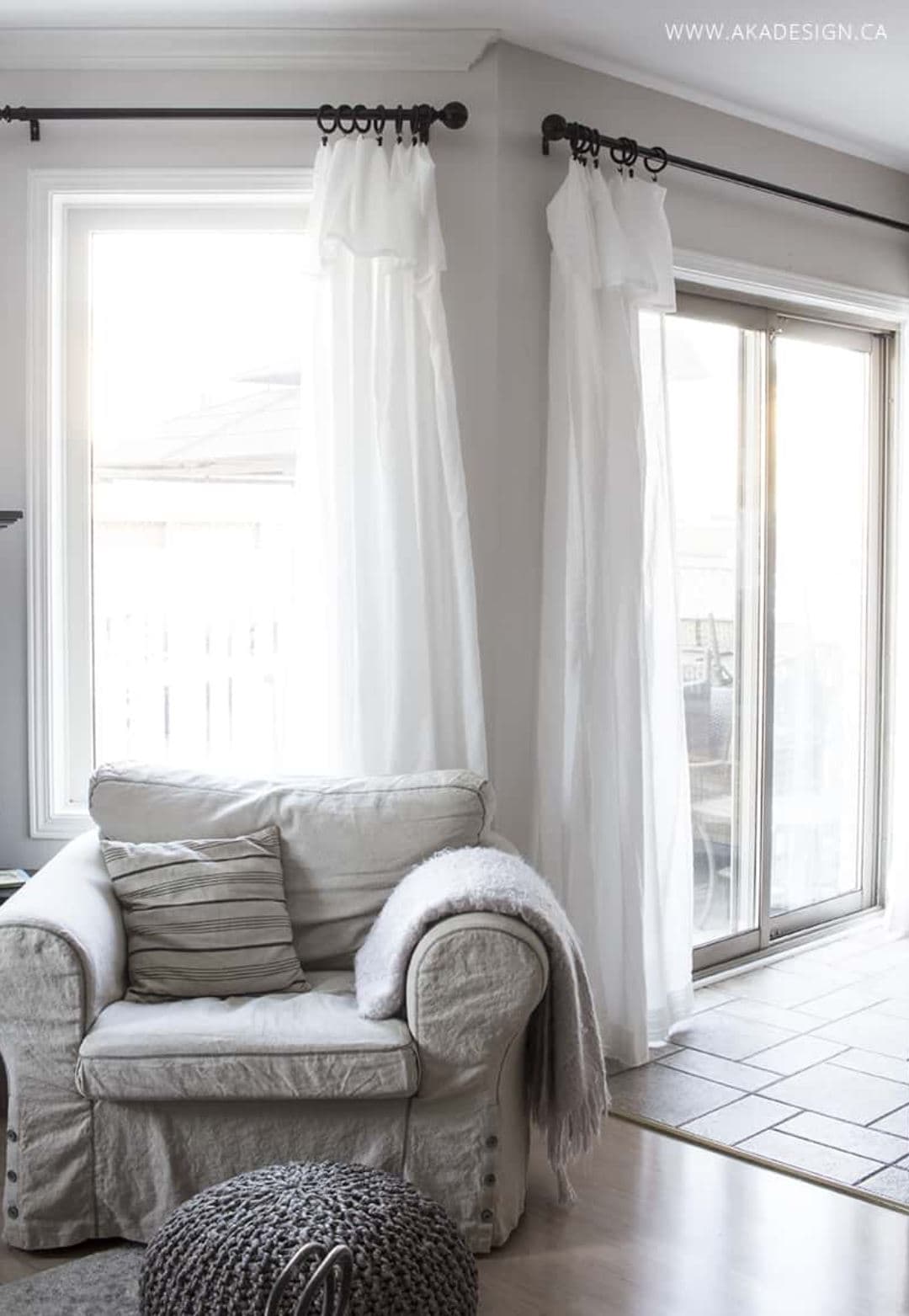 Ruffled white curtains hanging over large windows in a basement with a large gray armchair in front.