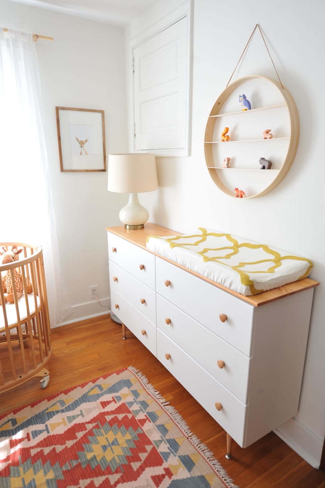 A white IKEA Tarva dresser with added legs and wooden knobs in a baby's nursery.