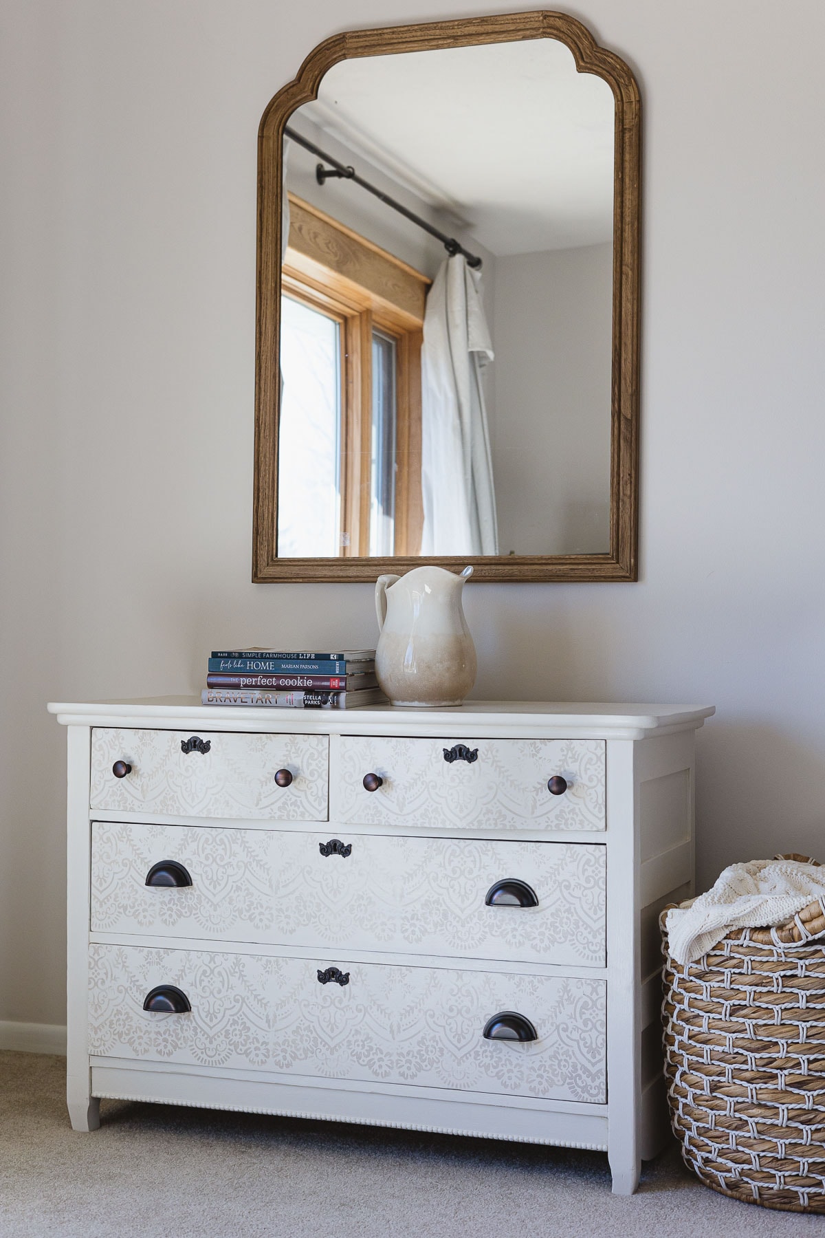 A painted dresser with a mirror hanging above, a stack of books and ironstone pitcher on top and a woven basket with blankets to the side.