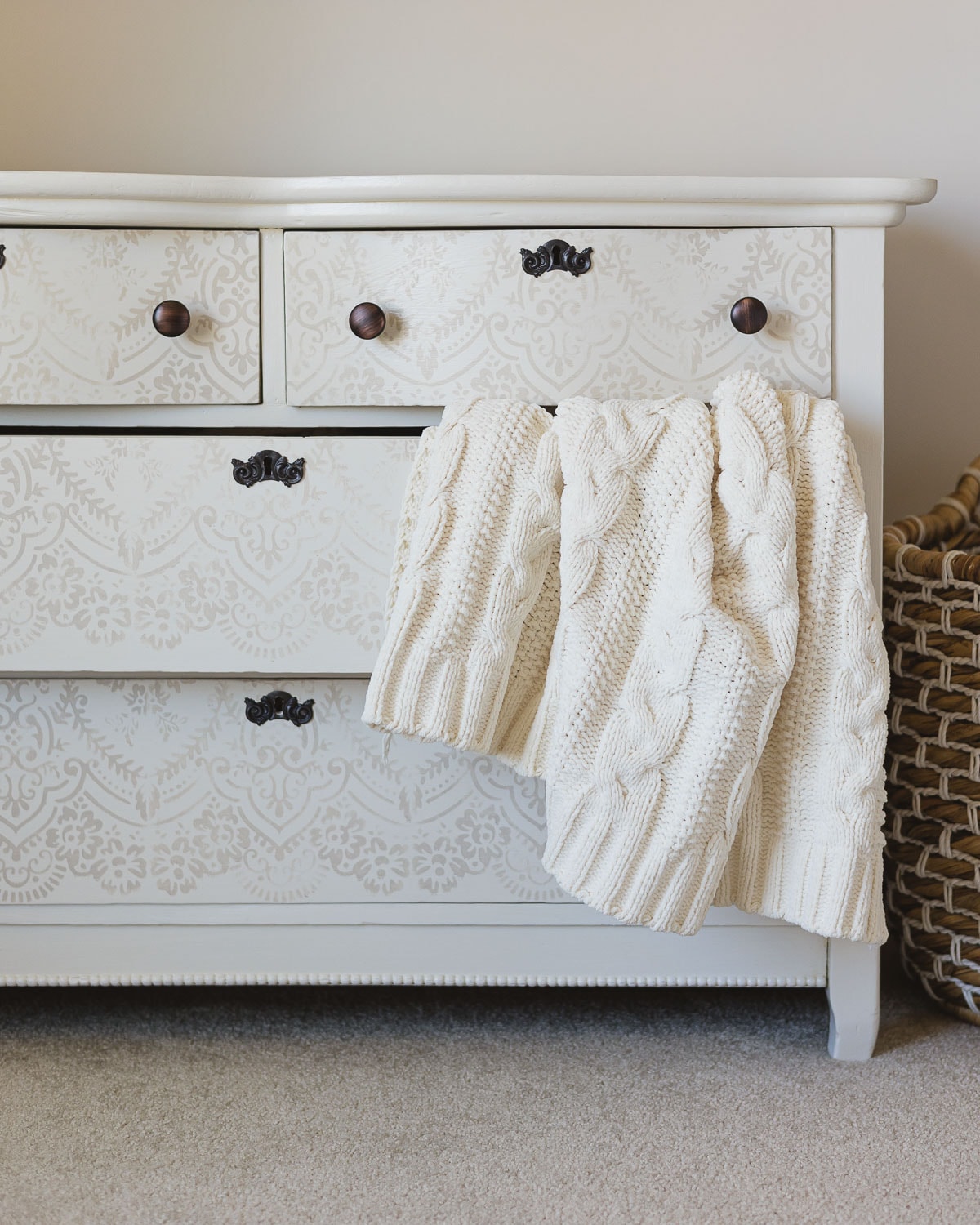 A white and taupe stenciled dresser with a thick creamy cabled blanket falling out of the middle drawer.