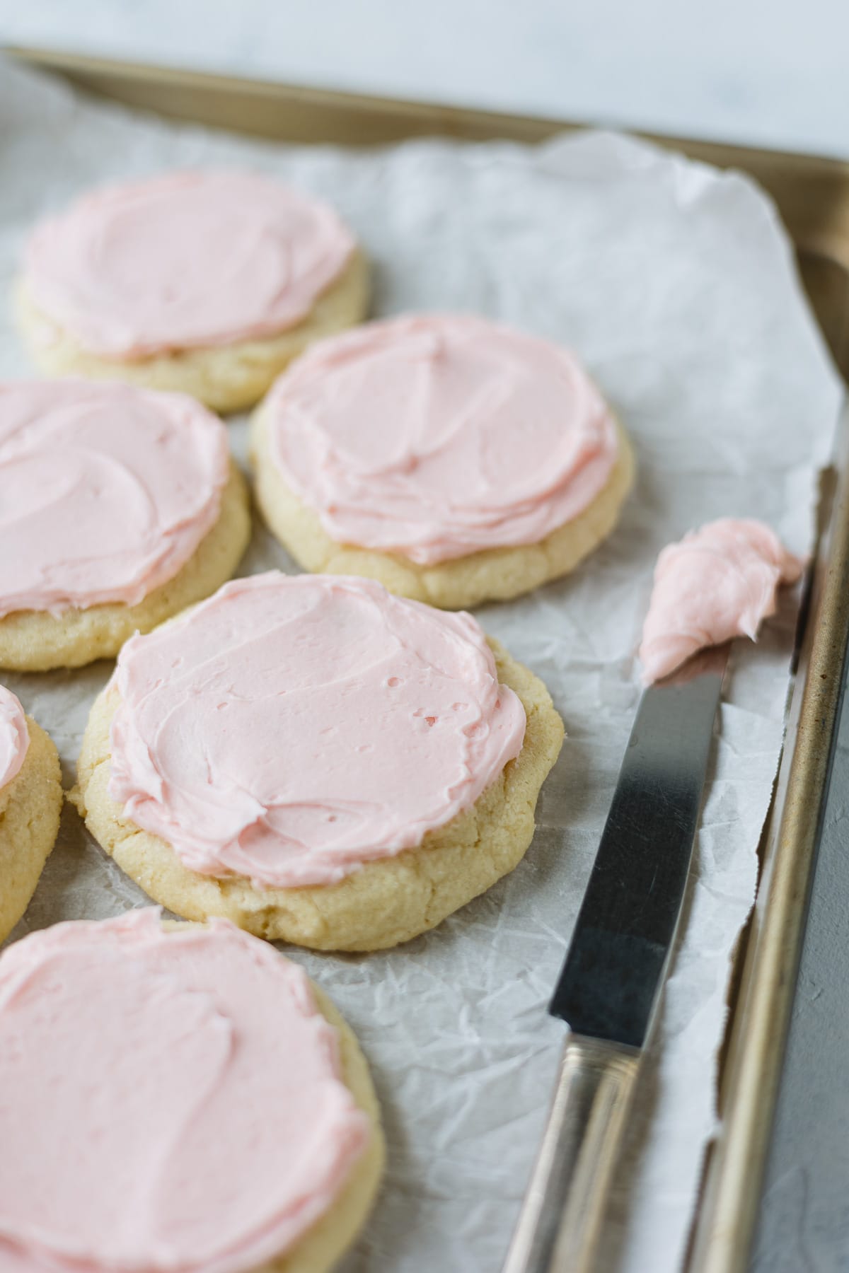 A parchment-lined cookie sheet filled with pink-frosted sugar cookies and a knife with frosting on it.