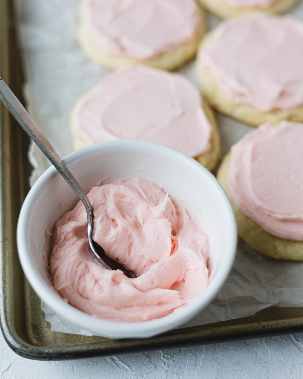 A small bowl filled with pink buttercream and a spoon on a cookie sheet.